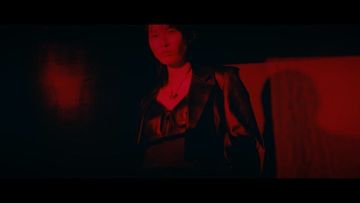 Adult female in black leather jacket stands against a red wall in a dark indoor setting with soft lighting.