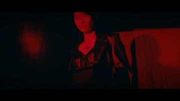 Adult female in black leather jacket stands against a red wall in a dark indoor setting with soft lighting.