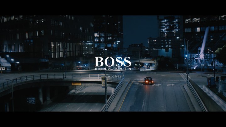 Night cityscape with a car driving on an empty road beneath an overpass, with the illuminated BOSS logo in the center of the frame.