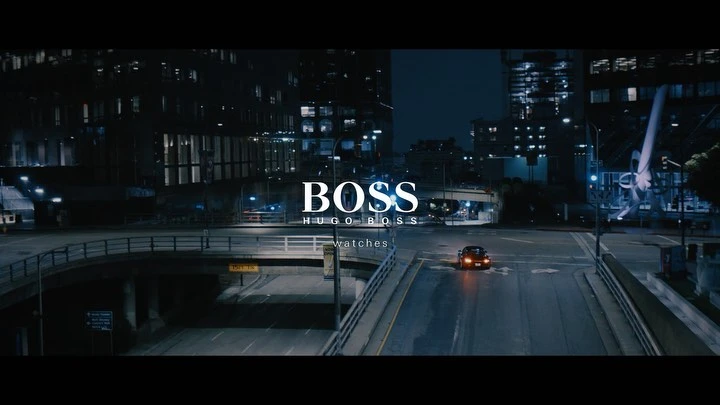 Night cityscape with a car driving on an empty road beneath an overpass, with the illuminated BOSS logo in the center of the frame.