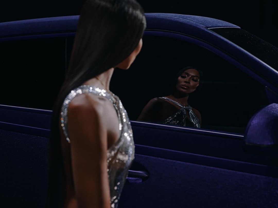 Fashionable adult women pose near a luxurious purple car in a studio, showcasing elegant dresses and sophisticated style in a dark, moody setting.