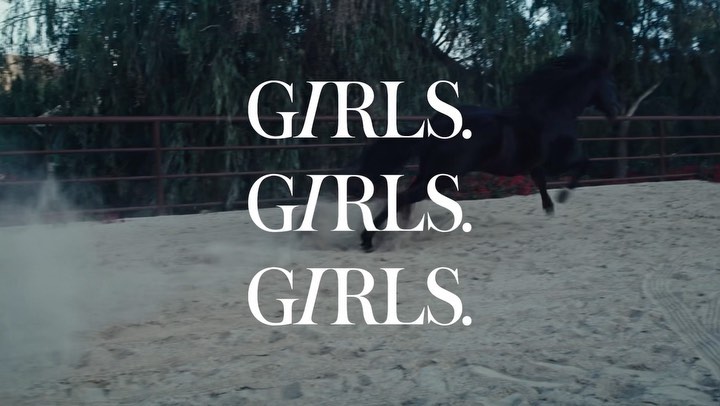 Dramatic shot of a black horse galloping in a sandy outdoor arena, framed by dark trees and overlaid with bold white text reading 'GIRLS'.