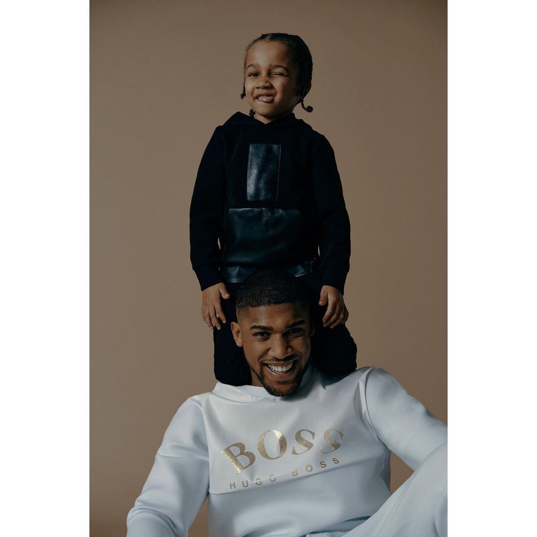 A Black father and his child pose in a studio; the child sits on the father's shoulders, wearing hoodies. The backdrop is plain.