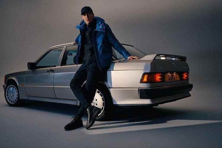 An adult male poses next to a vintage silver Mercedes-Benz car, wearing a dark cap, black pants, and a blue puffer jacket.