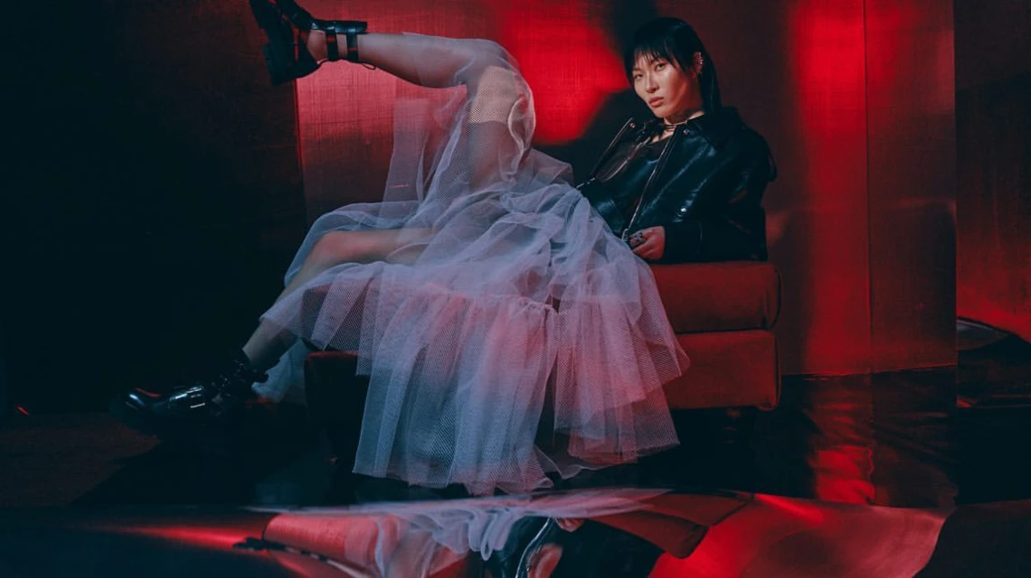 Stylish woman in black leather jacket and white tulle dress sits against a red wall in a dark room with dramatic lighting.