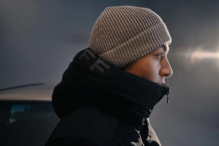 Profile shot of a man in a beanie and black jacket looking to the right against a blurred neutral background with light flare.