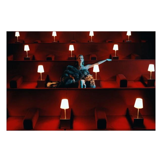 Elegant woman in gown reclines in an auditorium-style seating arrangement illuminated by table lamps against a red backdrop.