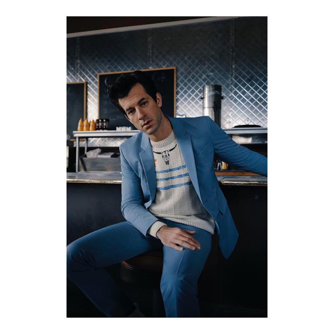 Stylish man in a light blue suit and patterned sweater seated in a diner, leaning against the counter in a medium shot.