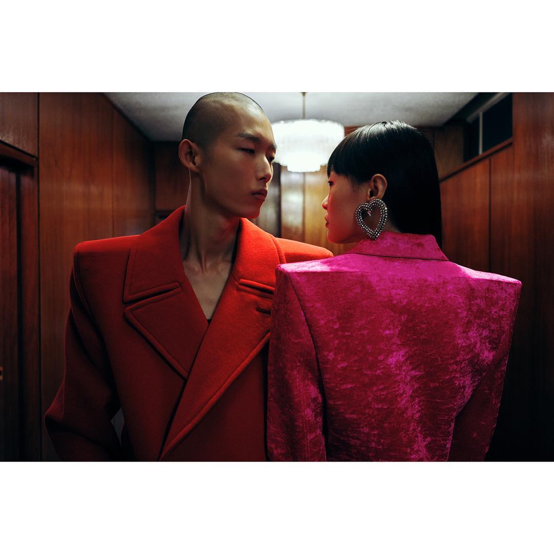 Two fashion models in bright overcoats pose indoors. A man in orange faces a woman in pink, showcasing oversized heart earrings and strong shoulder silhouettes.
