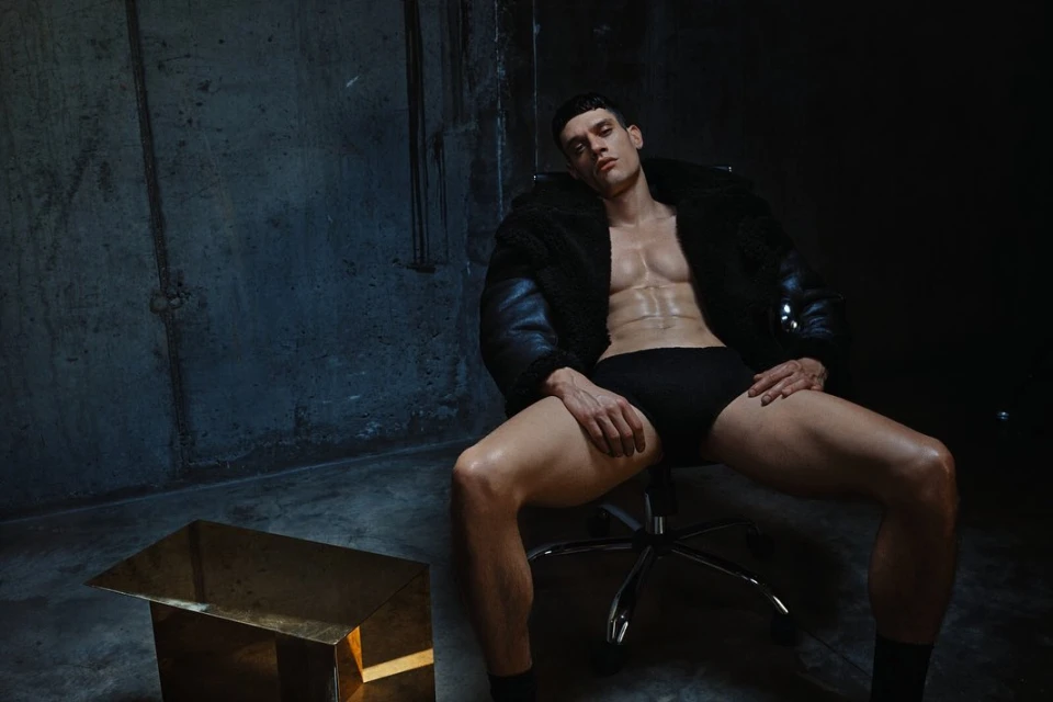 Portrait of a male fashion model in black underwear and a jacket sitting on an office chair in a dimly lit, industrial setting.