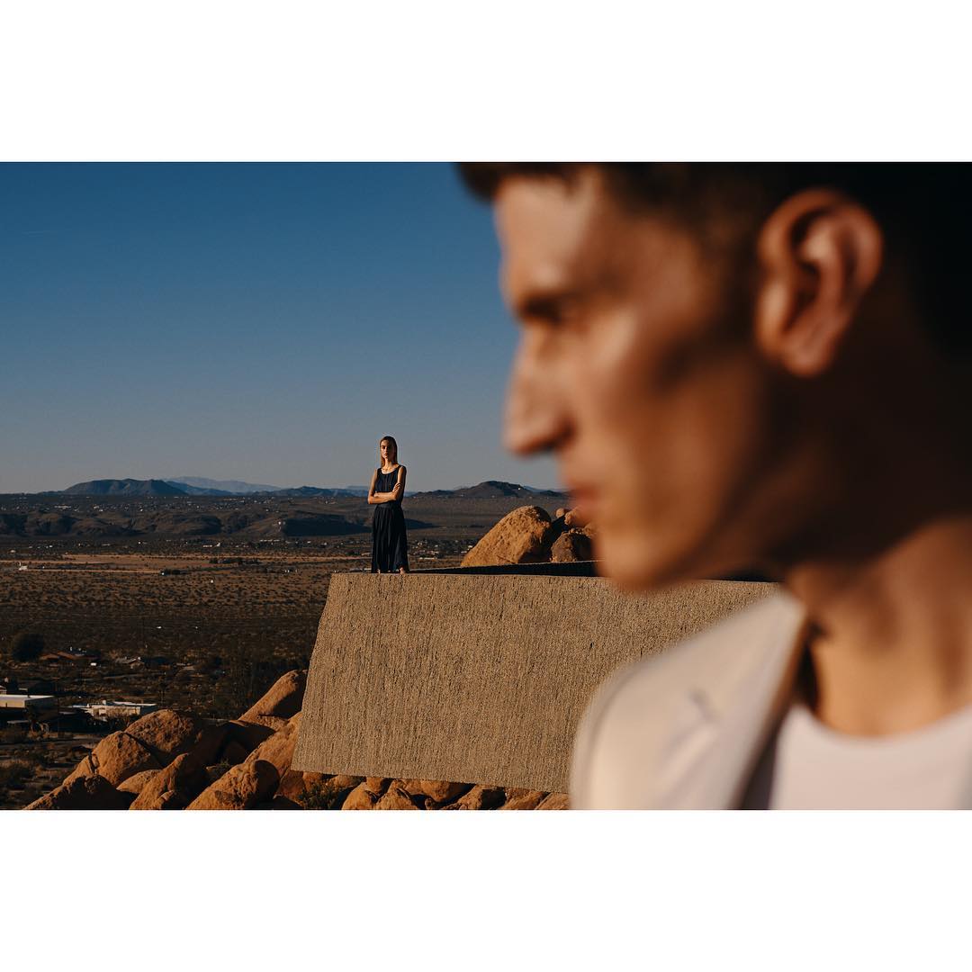 A man in a suit is blurred in the foreground as a woman in a dress stands on a ledge overlooking a desert landscape under a blue sky.