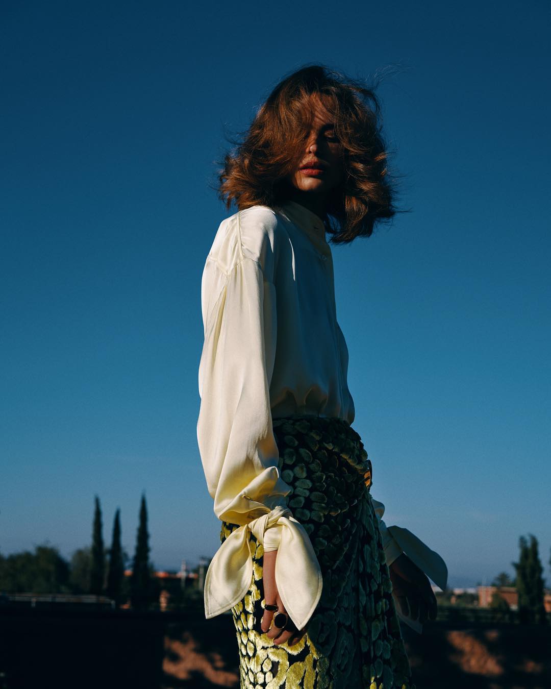 Portrait of a young adult woman in a white satin blouse and decorative wrap skirt posing outdoors against a blue sky backdrop.