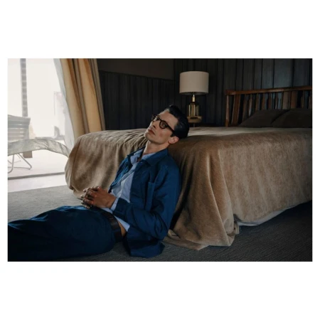 Man in blue shirt and trousers leaning back against a hotel bed in a room with a lamp and large window