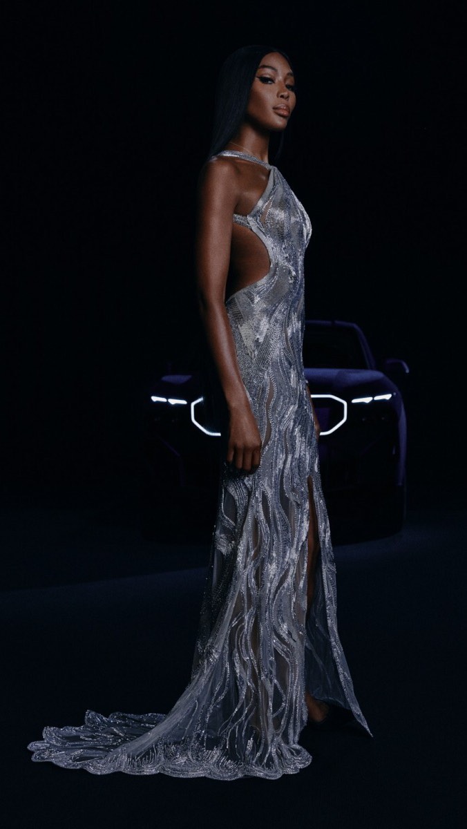 Elegant Black woman poses in shimmering dress against a dark backdrop with a blurred car detail, combining high fashion with automotive.