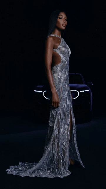 Elegant Black woman poses in shimmering dress against a dark backdrop with a blurred car detail, combining high fashion with automotive.