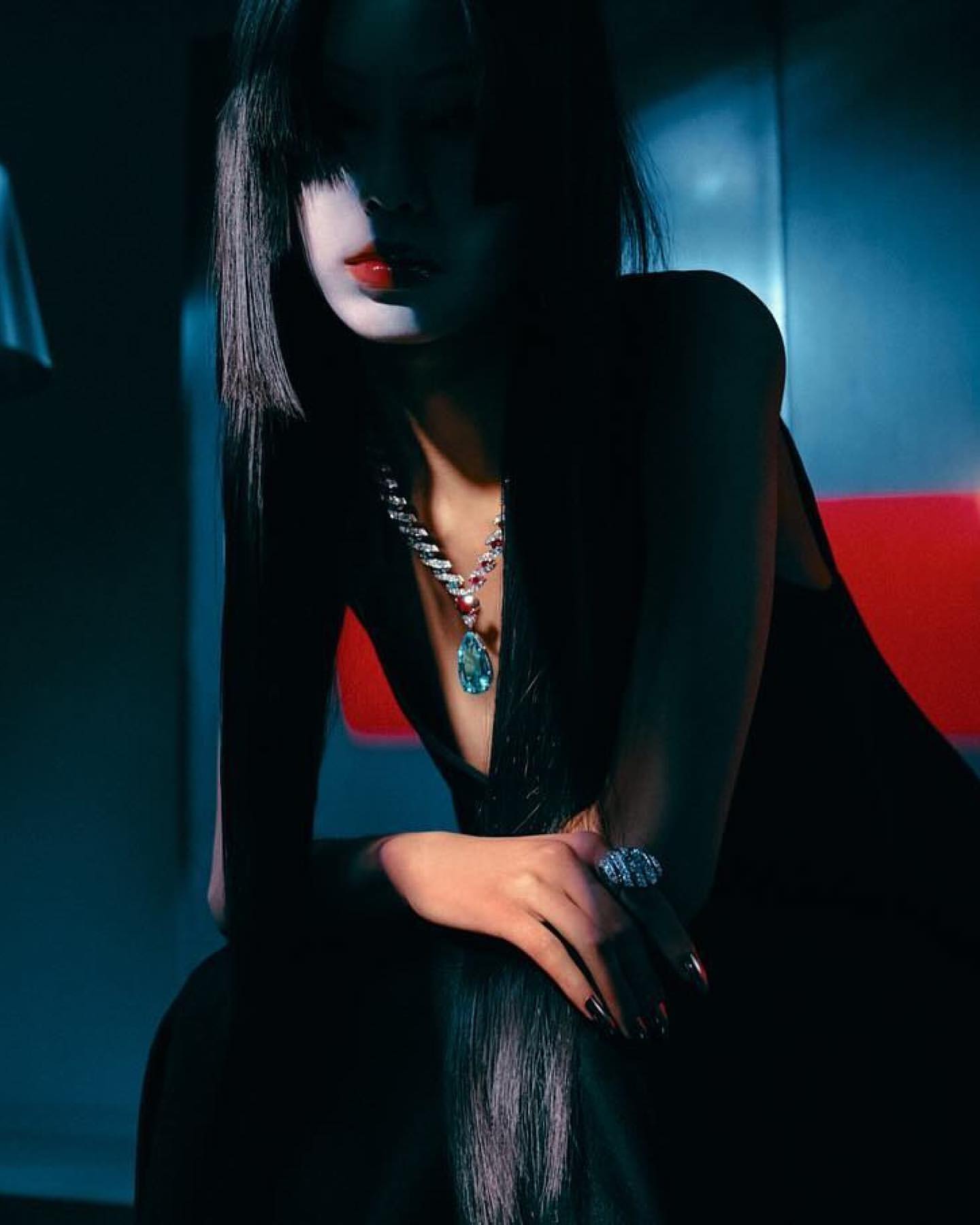 Woman with dark hair and red lipstick sits indoors wearing a black dress, diamond necklace, and ring, captured in moody lighting.