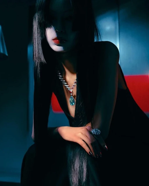 Woman with dark hair and red lipstick sits indoors wearing a black dress, diamond necklace, and ring, captured in moody lighting.