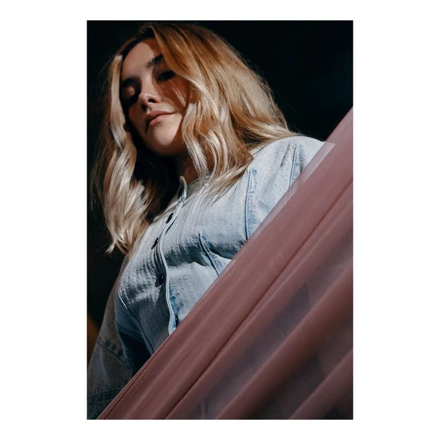 Blonde woman in a denim shirt poses in a low angle shot, with pink fabric draped in the foreground and a dark backdrop.
