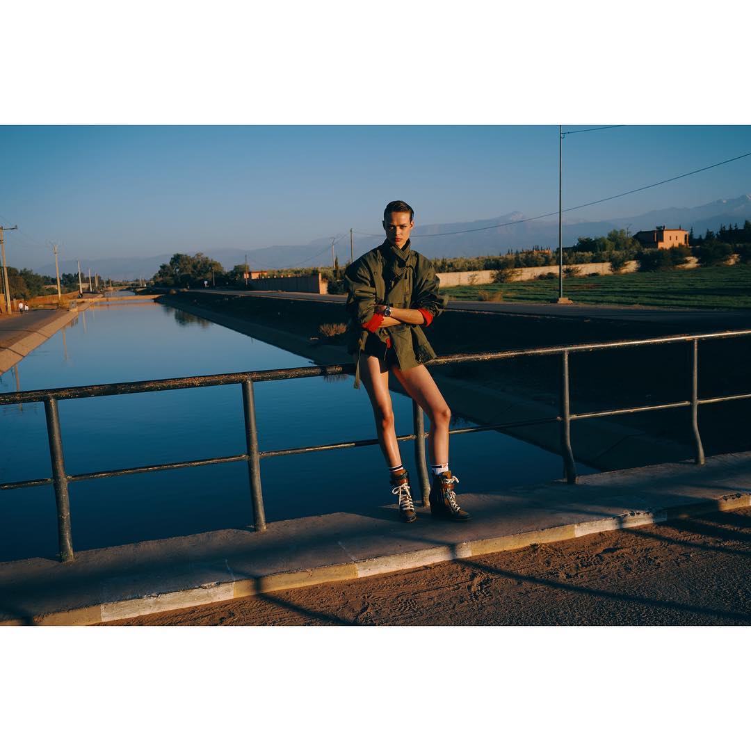 A person in a jacket poses on a bridge over a canal, wearing boots and socks, with mountains in the background on a sunny day.