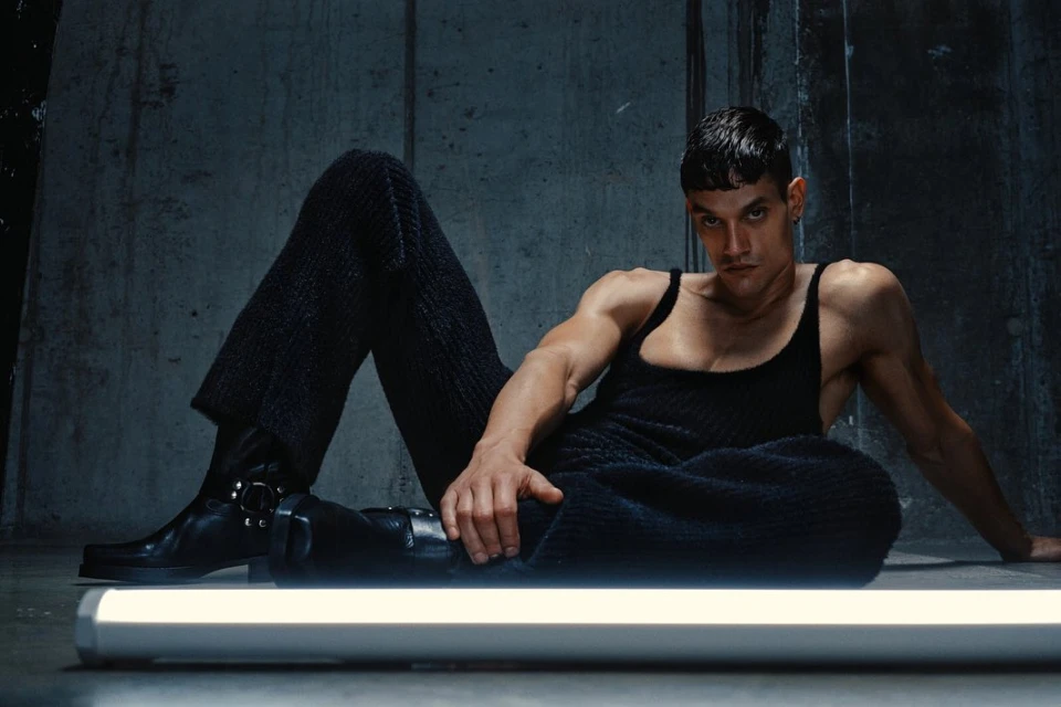 Athletic adult male laying indoors wearing a tank top and textured pants with combat boots, illuminated by a light fixture.
