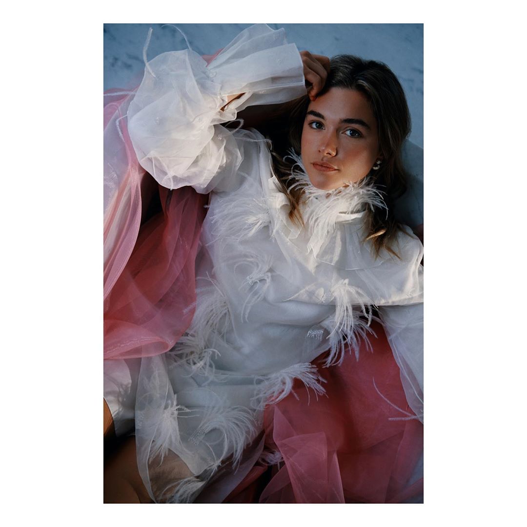 Portrait of a young woman lying down wearing a white, feathered dress and surrounded by pink tulle fabric in a studio setting.