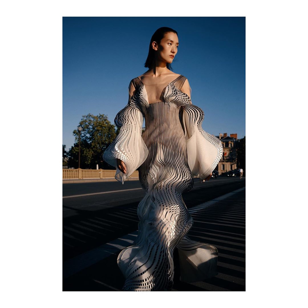 Model in a sculptural striped optical illusion dress standing outdoors against a bridge and blue sky