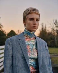 A person with short blonde hair wearing a plaid blazer and a colorful sequin turtleneck in an outdoor setting at dusk.