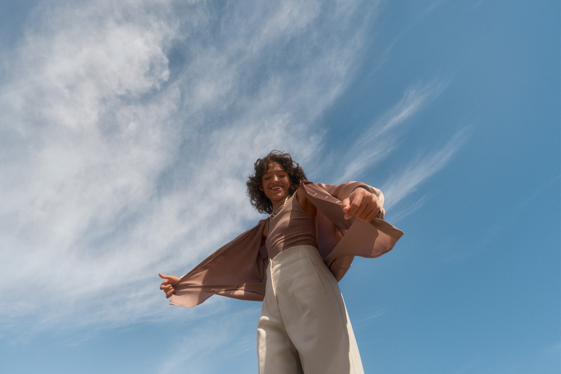 Adult female in a mauve top and light pants smiles at the camera with arms out. A blue sky with light clouds provides a cheerful backdrop.