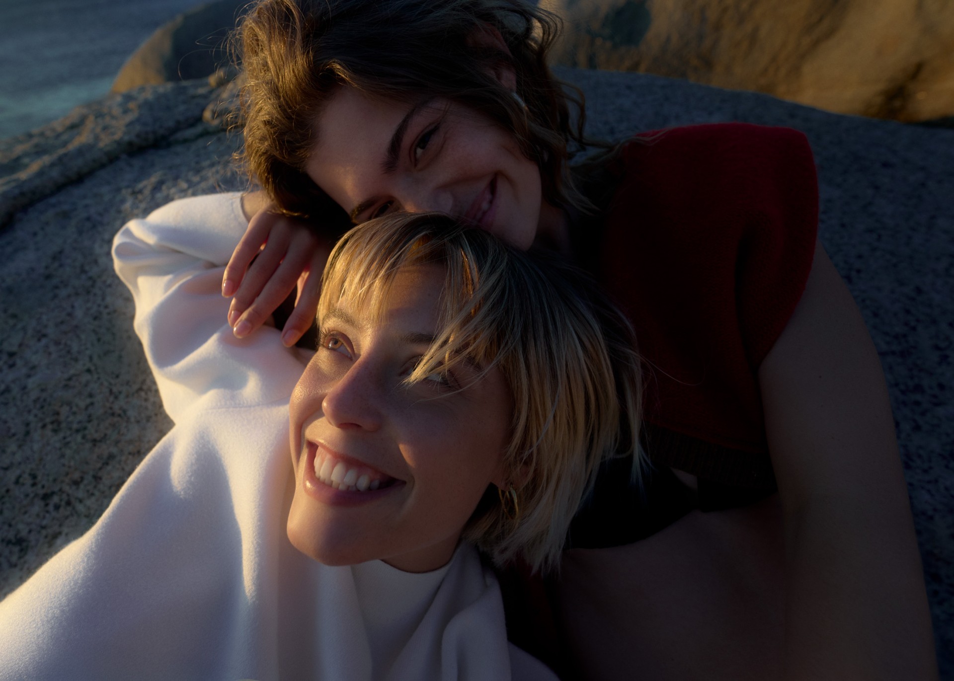 Two women, one in a red shirt and the other in a white hoodie, smiling and posing against a textured rock surface in natural light.