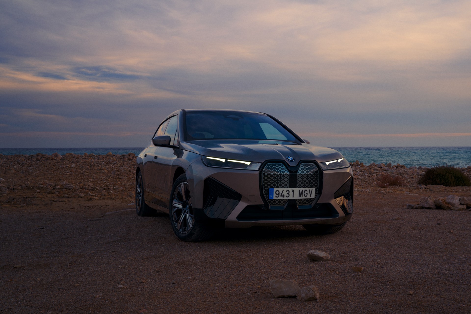 Modern grey BMW iX electric SUV parked on a rocky beach at sunset, with ocean and sky visible in the background. Vehicle front view.