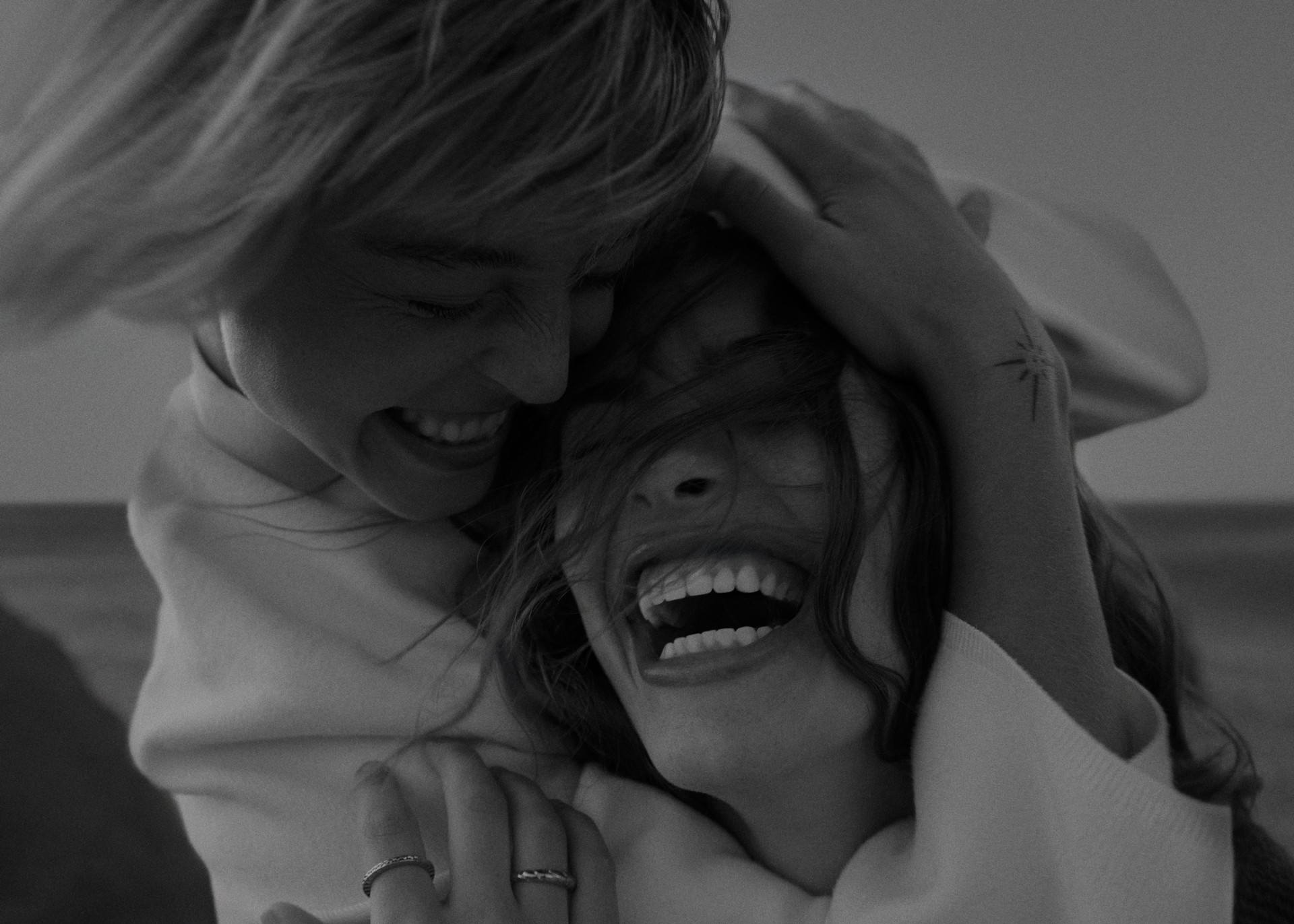 Two women embrace outdoors in black and white. One woman's face is visible, laughing; the other has her hands on her friend's head.