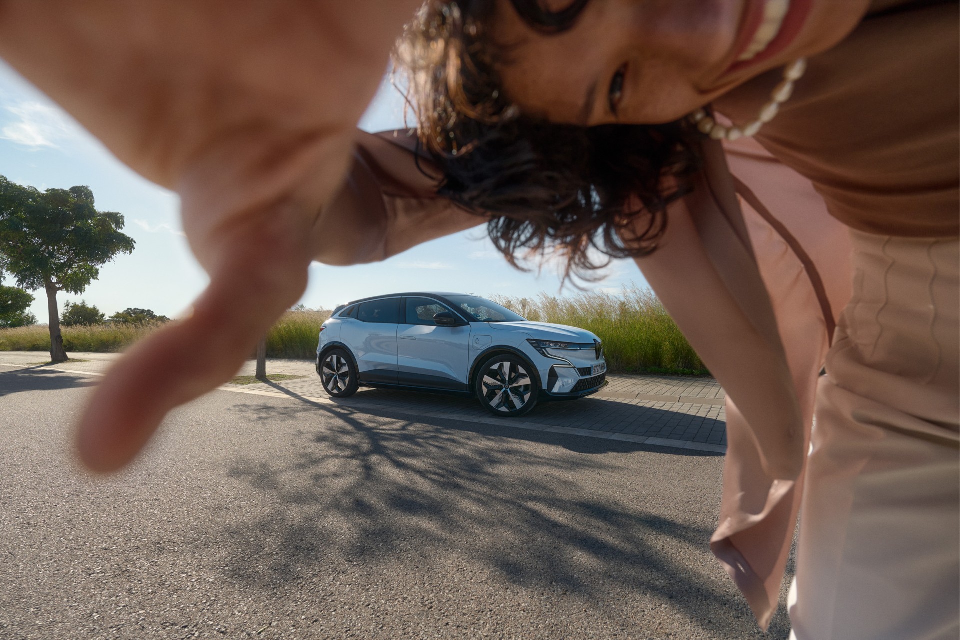 Light blue car parked near green grassy field with blue sky. Adult female partially visible taking a photo of vehicle outdoors.