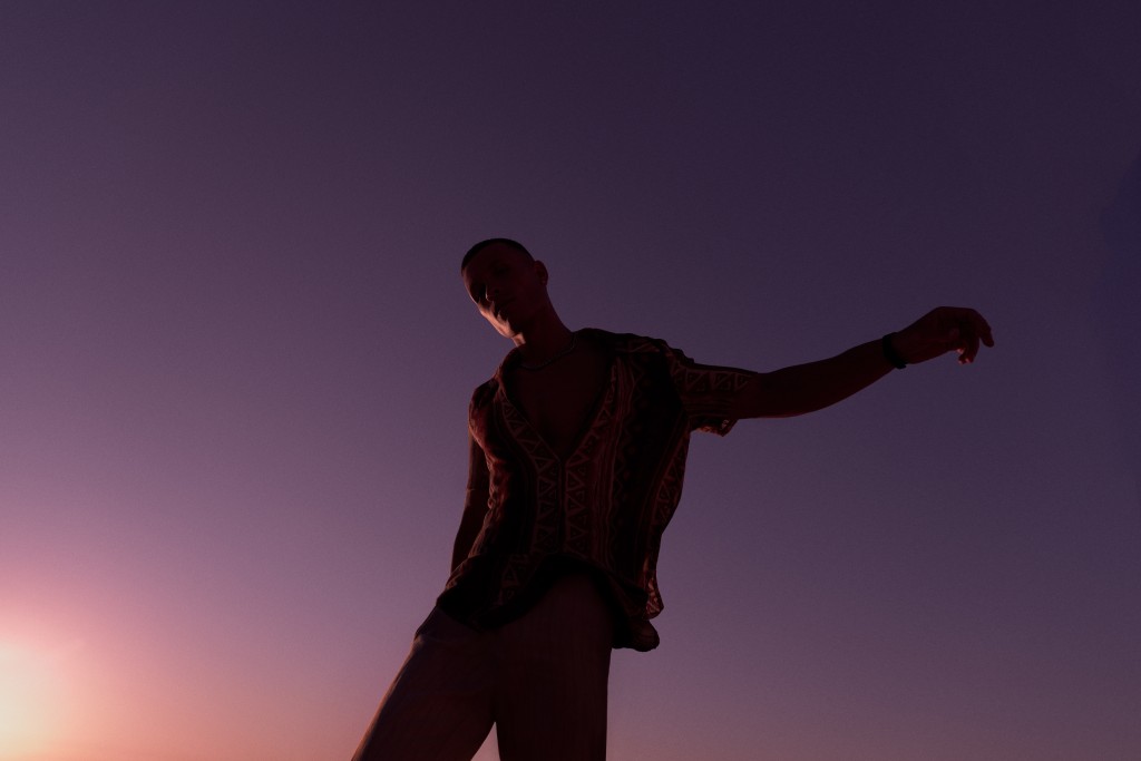 Man silhouetted against a purple sky wearing an open, patterned shirt, arm outstretched, casual pose, dusk lighting, low angle.