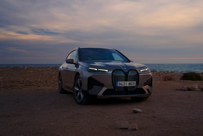 Modern luxury SUV with illuminated headlights parked on a rocky shore overlooking the ocean at dusk.