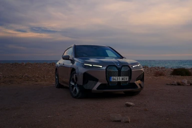 Modern luxury SUV with illuminated headlights parked on a rocky shore overlooking the ocean at dusk.