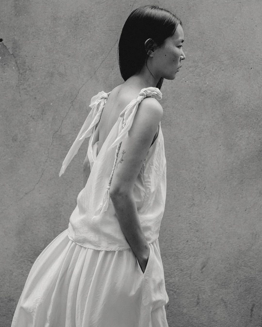 Monochrome portrait of an Asian woman in a white dress against a concrete wall, showcasing fashion and editorial style in natural light.