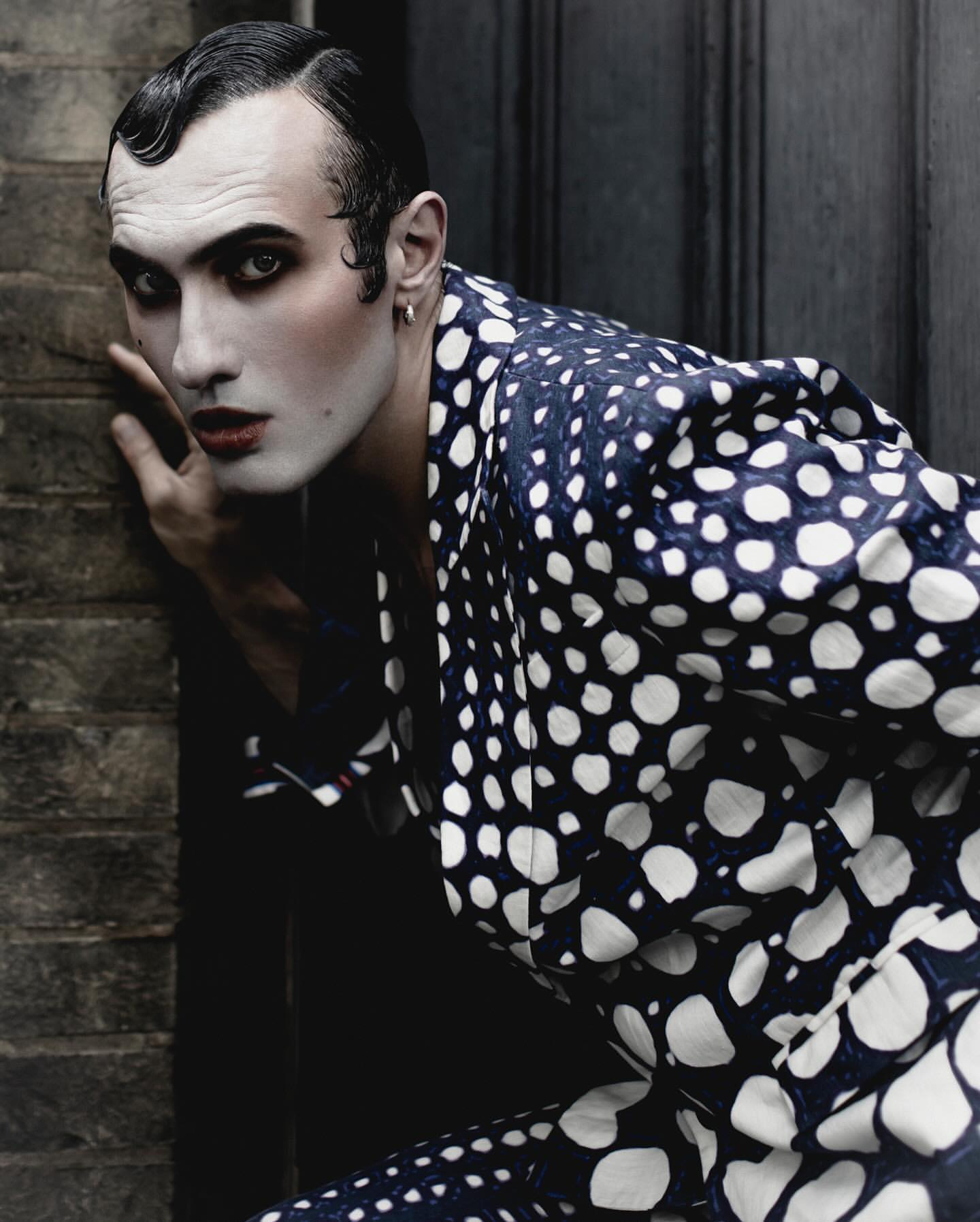 A striking fashion portrait of a person in theatrical white makeup and a bold polka-dot jacket poses near a textured stone wall.