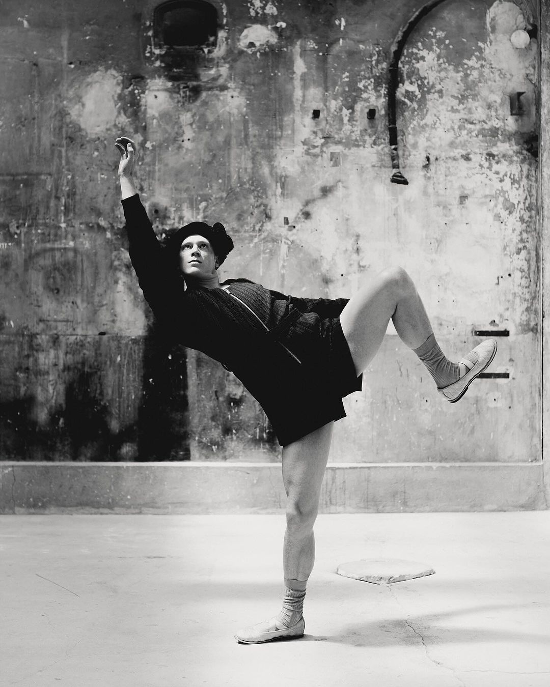 Male ballet dancer strikes a pose mid-performance, balancing on one leg in front of a distressed wall inside a studio.