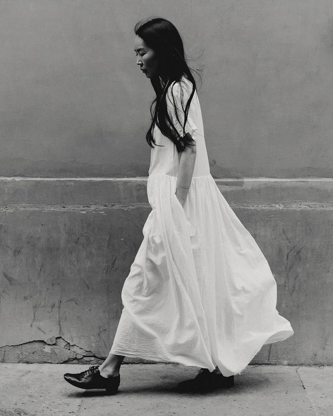 An Asian woman in a white dress and black shoes walking against a textured wall in a black and white outdoor fashion shot.