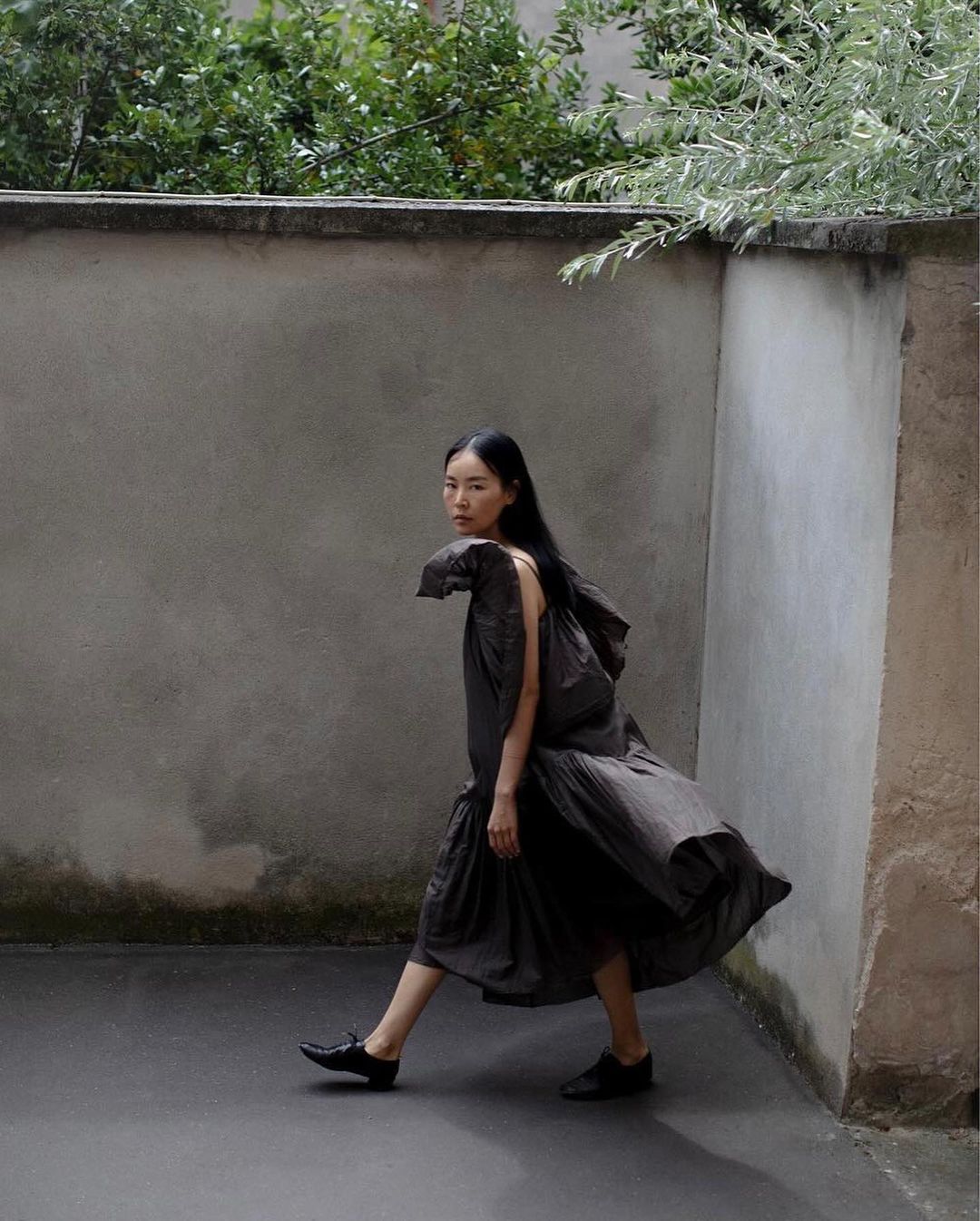An Asian adult woman in a dark flowing dress is walking in a concrete alley with green foliage visible overhead outdoors in daytime.