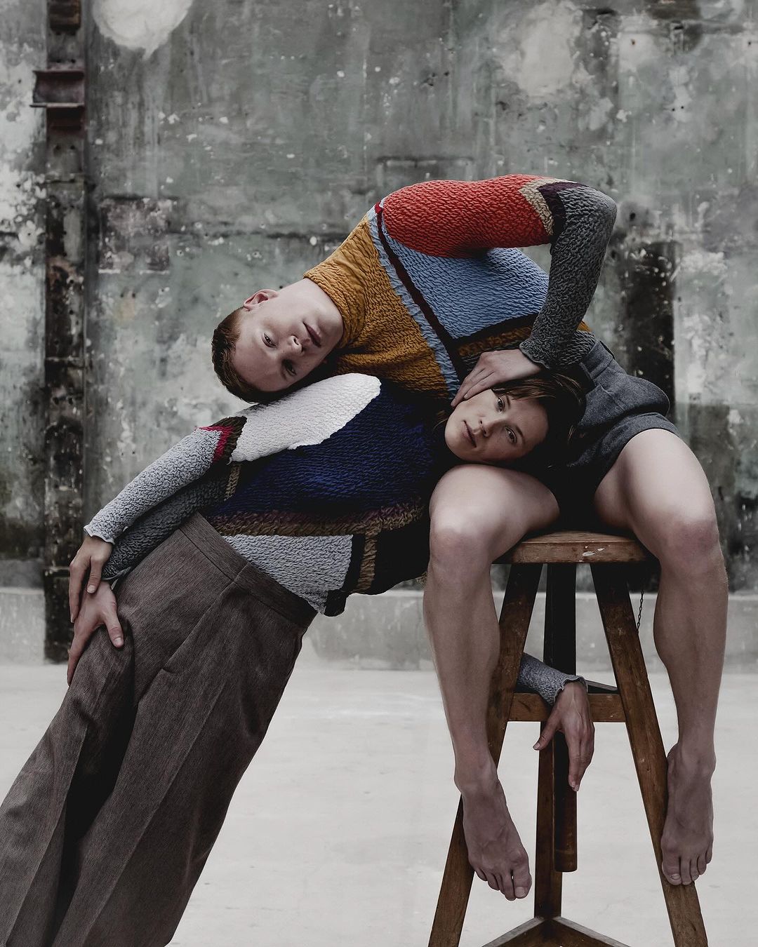 Two fair-skinned adults in sweaters creatively pose in a worn studio with gray floors and walls, one seated on a stool with bare feet.