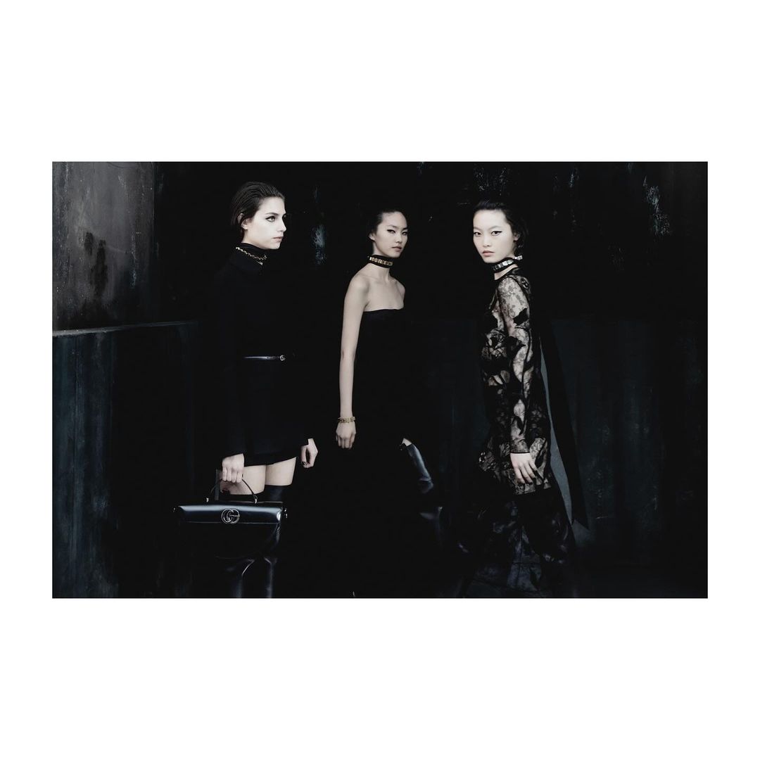 Three women in black outfits posing against a dark background, wearing choker necklaces and a black purse, high fashion photography.