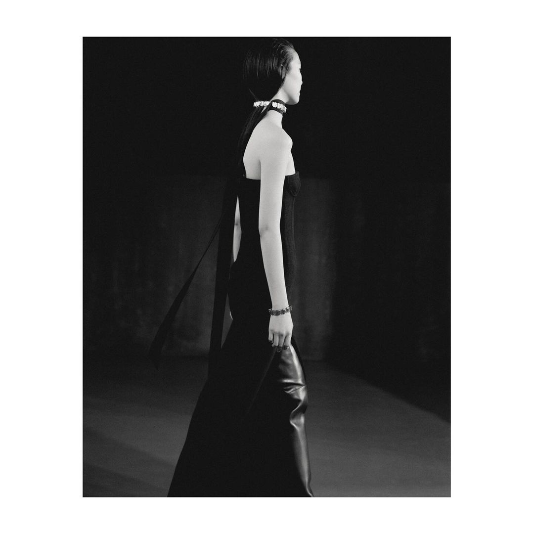 A side-profile shot of a fashion model in a black dress and pearl accessories, set against a dark backdrop in a photo studio.