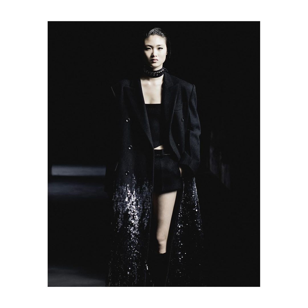 A woman in a black outfit and long coat poses in a dimly lit studio with a dark background. Fashion-forward apparel.