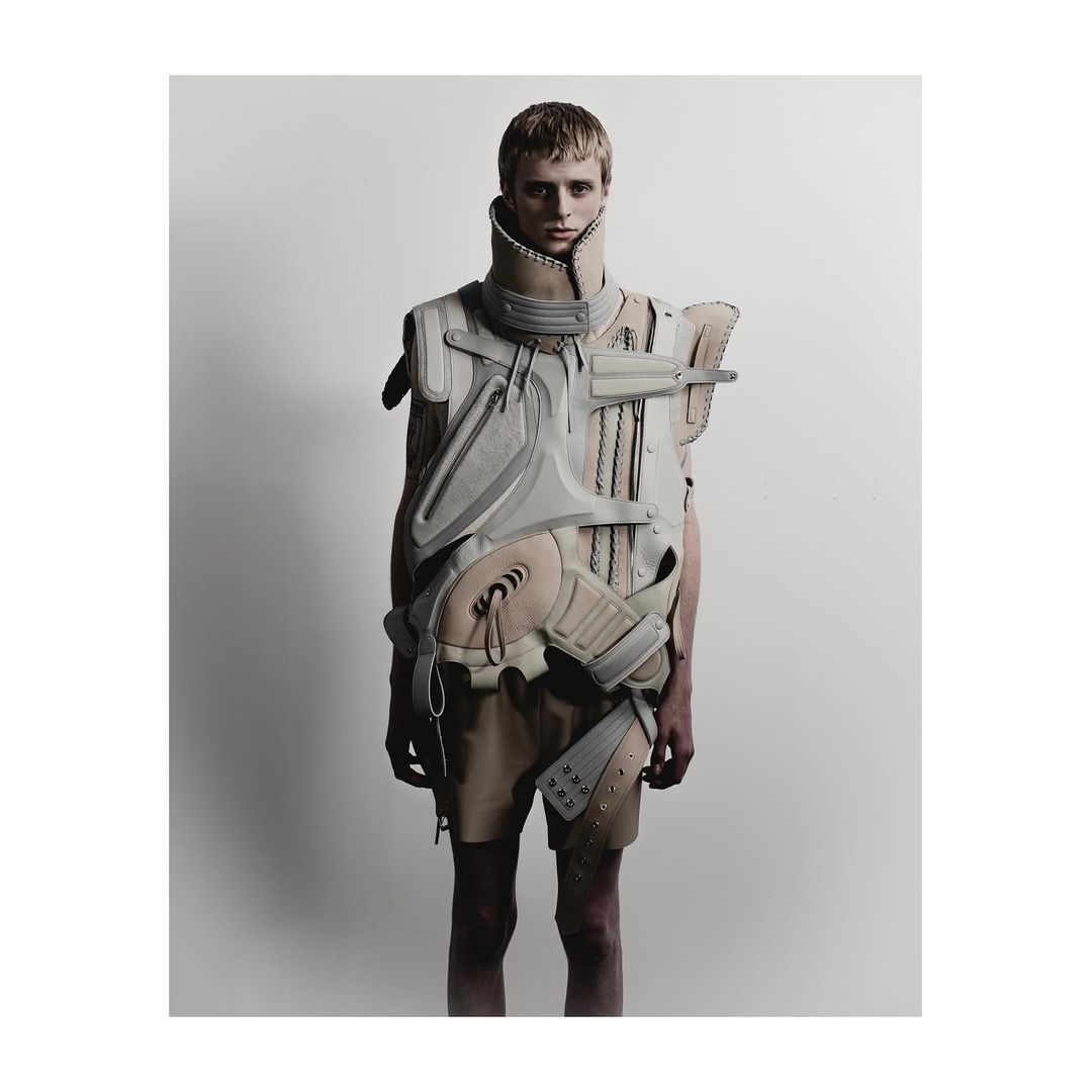 Full-length portrait of a fair-skinned, blond adult male wearing futuristic armor in a studio with a neutral background.