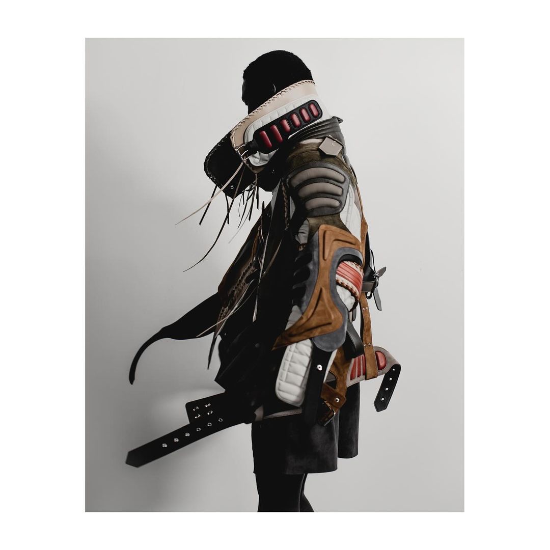 Person in avant-garde fashion posing against a white wall. Unique garment made of layered materials, accessories, and black accents.
