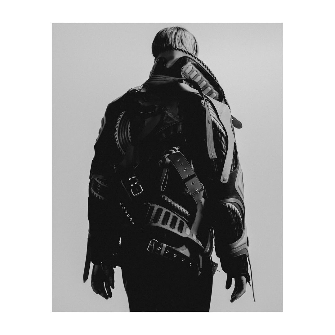 Monochrome back shot of person wearing an avant-garde leather-like jacket with straps and buckles against a neutral studio backdrop.
