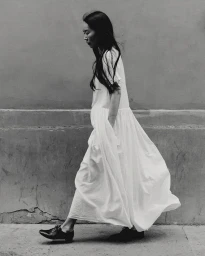 Monochrome portrait of a woman in a long white dress walking past a textured wall in Paris by Paul Phung.