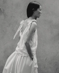 Monochrome portrait of an Asian woman in a sleeveless white top with tied shoulder straps against a textured wall