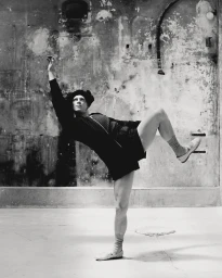 Dancer in dark athletic wear balancing on one leg in a dynamic pose against a weathered concrete wall in Paris.